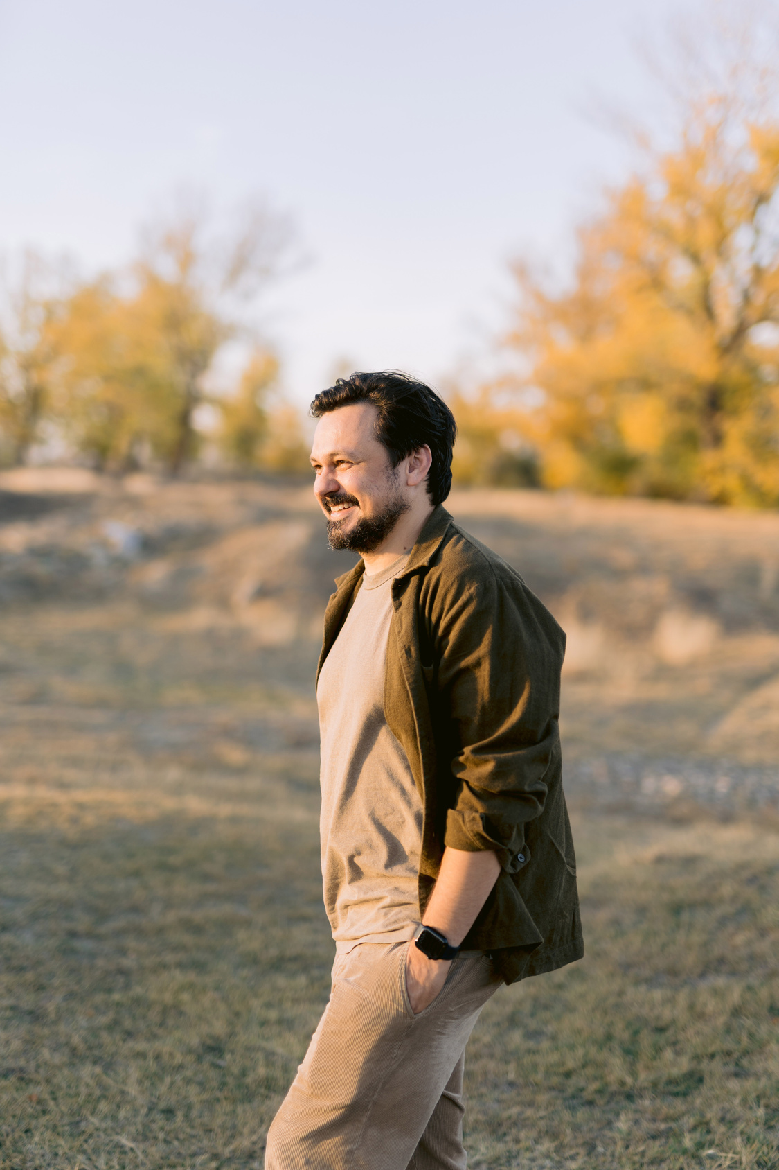 Man laughing outdoors in an autumn landscape wearing a green jacket over beige clothing