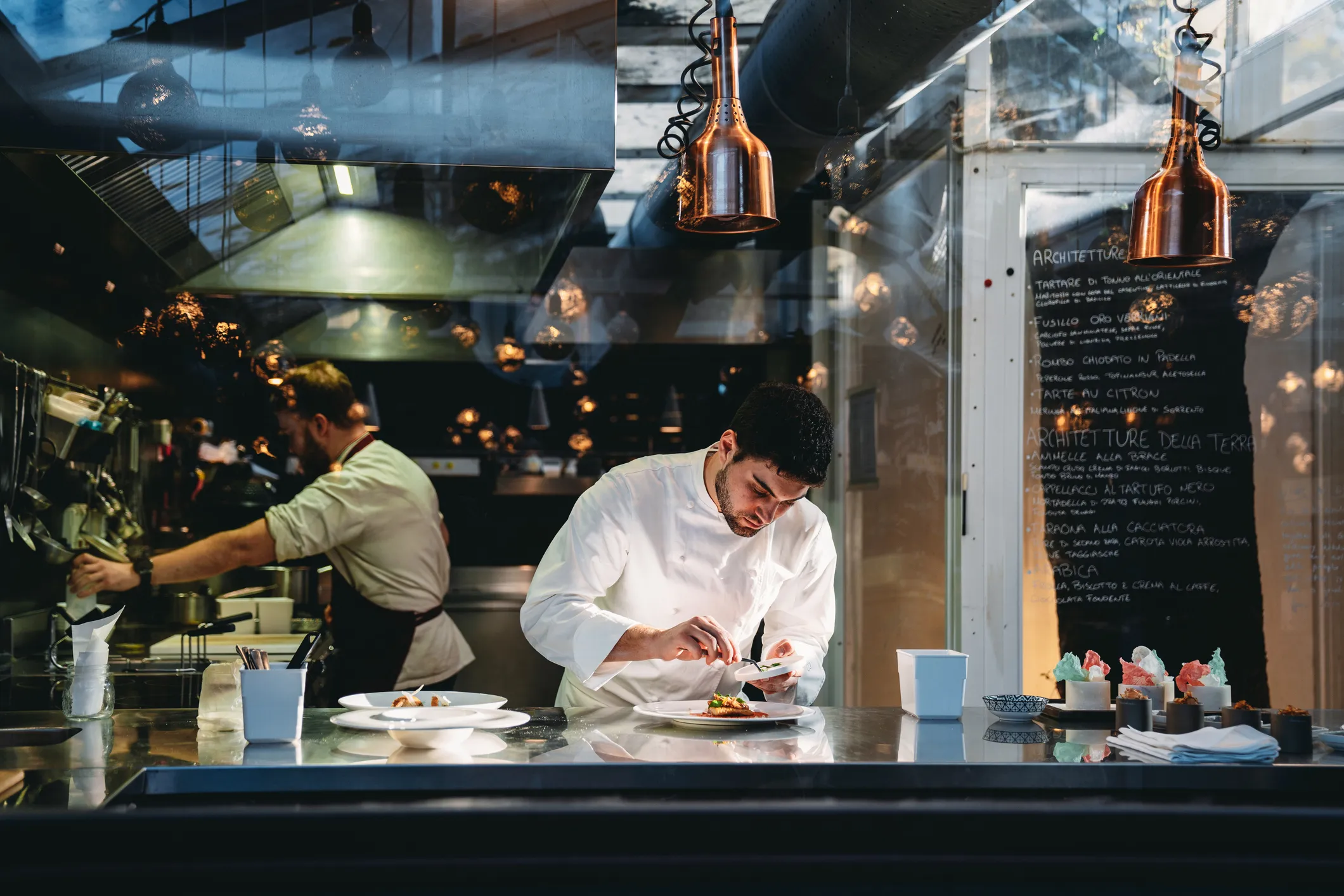 Two friends laughing and eating together at a restaurant counter with copper pendant lights overhead