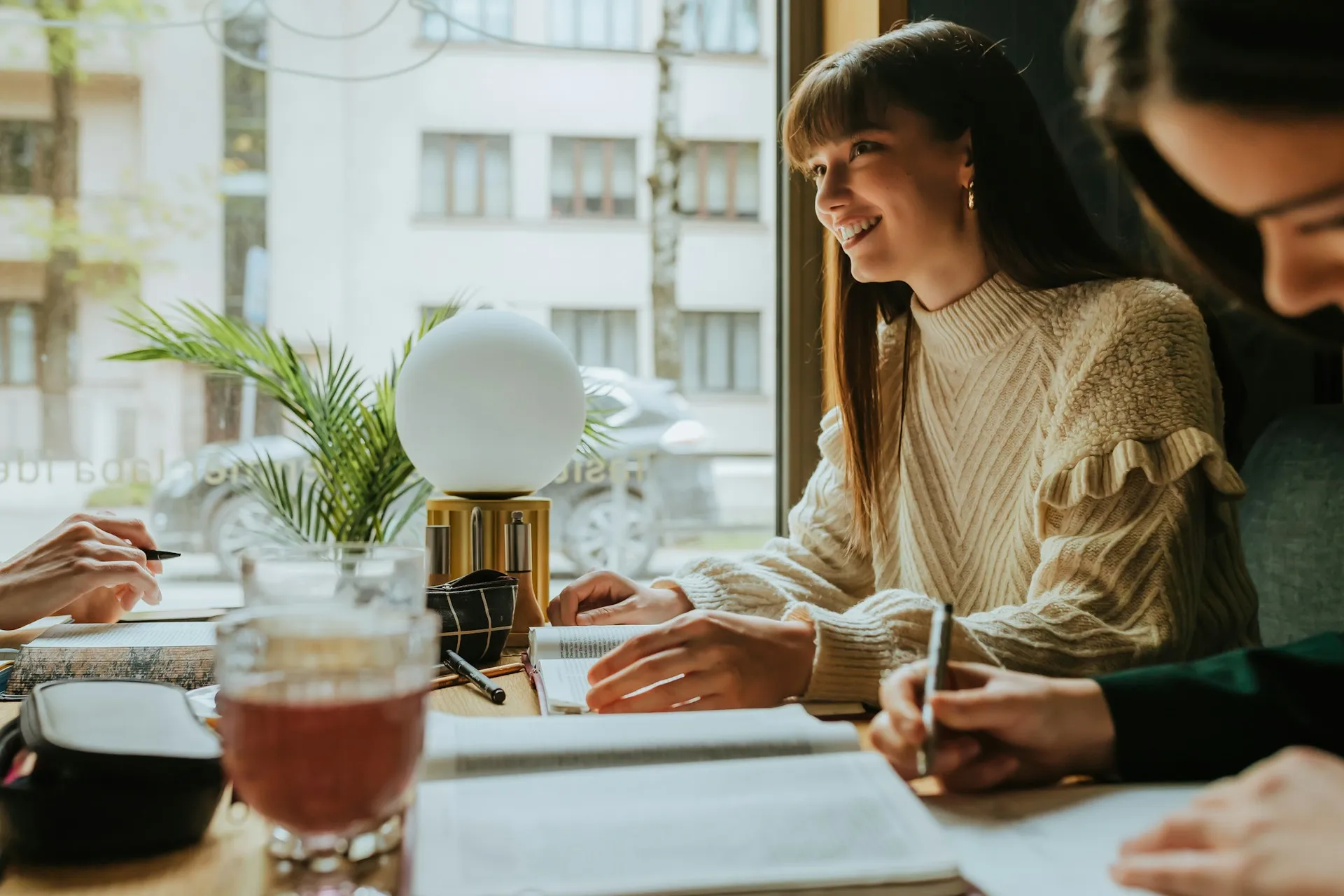 A woman smiling while writing in a notebook at a cafe table next to a large window.