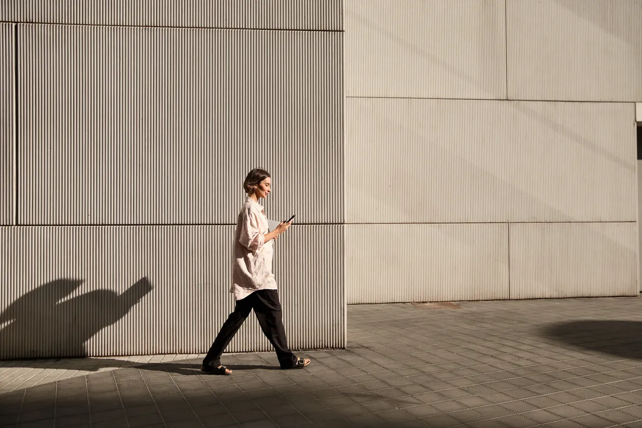 Person walking past a modern building using a phone.