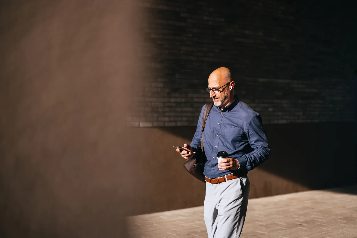 A man in a blue shirt and glasses walking outdoors while looking at his phone and holding a coffee cup.