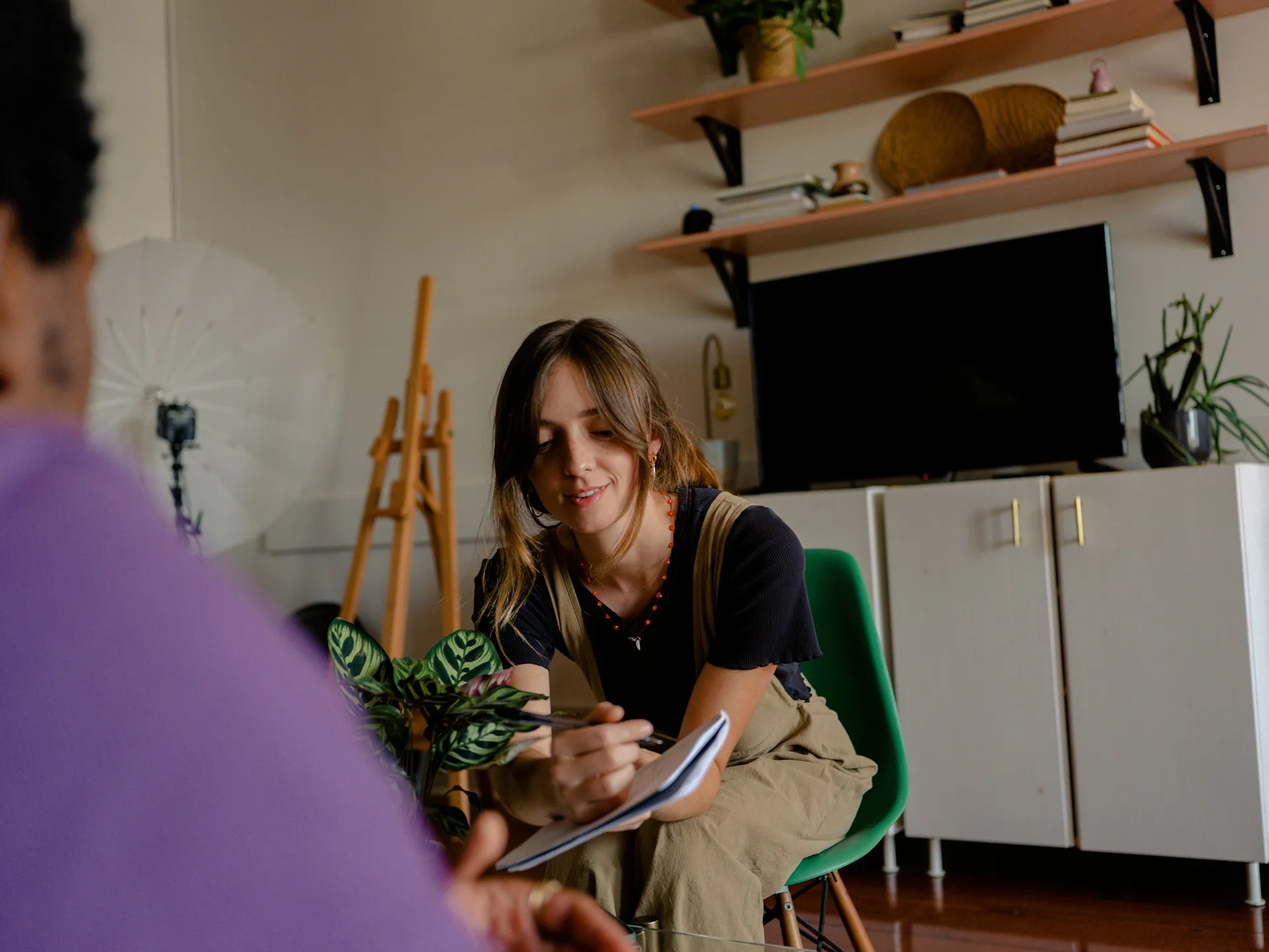 A woman sitting in a creative workspace, smiling while writing in a notebook.